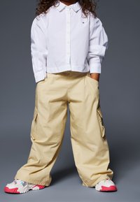 Kind met krullend haar, gekleed in een wit overhemd met knopen, beige wijde cargobroek met zakken en witte sneakers met roze zolen.