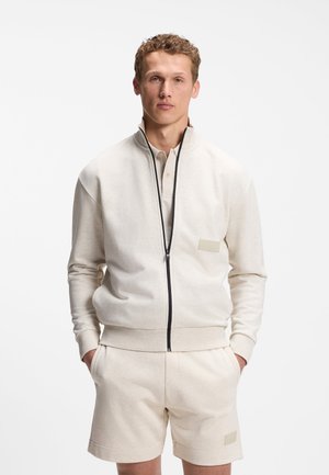 Mand med krøllet hår iført cremefarvet lynlåstrøje og matchende shorts, står med hænderne i lommerne mod en ensfarvet baggrund.