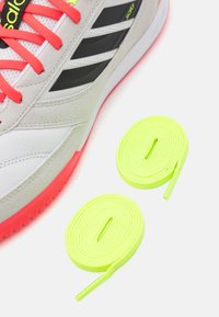 Zapatilla deportiva gris y blanca con acentos rojos y rayas negras, acompañada de dos cordones redondos de neón verde.
