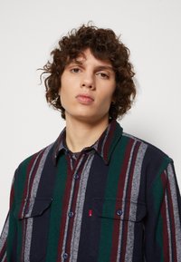 Camisa de franela de botones con rayas verticales en tonos azul marino, verde, burdeos y gris. Dos bolsillos en el pecho, diseño con cuello y tejido texturizado.