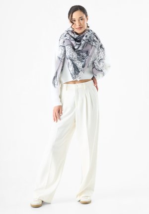 Oversized lichtgrijze sjaal met zwart dierenprint, gecombineerd met een witte crop-top trui en hooggetailleerde crèmekleurige broeken. Schoenen zijn witte sneakers.