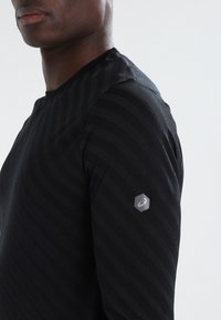 Musta pitkähihainen urheilupaita, jossa on hienovaraiset vaakasuorat raidat, pyöreä kaula-aukko ja kuusikulmainen logovivahde oikeassa hihassa.