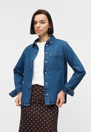 Ung kvinne iført en åpen blå denimjakke over en hvit topp og et brunt skjørt med prikkmønster, stående mot en ensfarget bakgrunn.