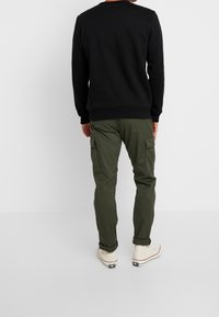 Sweat-shirt noir à col rond, associé à un pantalon cargo vert olive avec des poches latérales ; baskets blanches avec des touches rouges.