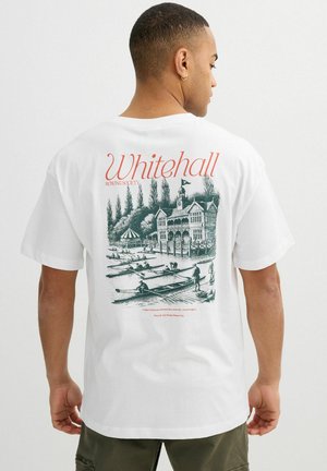 Hombre vistiendo camiseta blanca con escena detallada de club de remo y texto "Whitehall Rowing Society" en la espalda, de espaldas a la cámara.