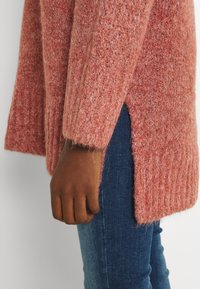 Dikke gebreide trui in zacht koraal met ribbeltextuur en zijsplitten, gecombineerd met donkerdenim jeans. Close-up van een arm en stofdetail.