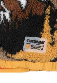 Fleece testurizzato marrone e nero con un bordo inferiore giallo; presenta un'etichetta del marchio Timberland con logo arancione e dettagli testuali.