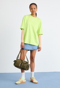 Neon zelené oversized tričko, svetlo béžové dlhé rukávy pod ním, denimová minisukňa, olivovo zelená taška s viacerými vreckami a pastelové tenisky s oranžovými pruhmi.