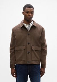 Giacca marrone in misto lana con chiusura a bottoni, due ampie tasche a soffietto e colletto, indossata sopra una camicia a righe. Design casual.