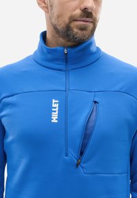 Mann trägt einen leuchtend blauen Reißverschluss-Pullover mit "MILLET"-Logo und teilweise geöffnetem Brusttasche, die ein blaues Netzfutter zeigt.