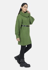 Robe-pull verte tricotée avec un col montant et une texture côtelée, ceinturée avec une ceinture noire. Portée avec des leggings noirs et des bottes épaisses noires.