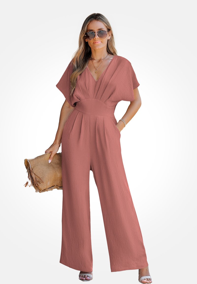 Une combinaison rose corail avec une finition texturée, des jambes larges et un décolleté en V profond. Le mannequin tient une pochette à franges et porte des lunettes de soleil.