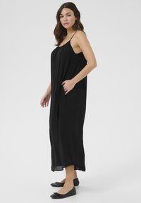 Vestido largo negro sin mangas de corte suelto, con detalle de bolsillos y fabricado en una tela ligera. Combinado con zapatos planos negros.