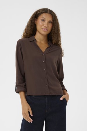 Brown button-up shirt with a collar and rolled sleeves, made from smooth fabric. Paired with dark denim pants. No patterns or accents visible.