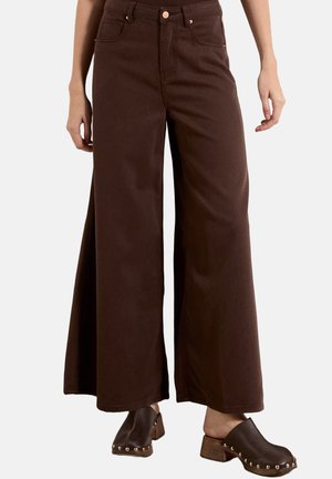 Donna con pantaloni marroni a vita alta e gamba larga abbinati a zoccoli marrone scuro con tacco in legno e borchie metalliche.