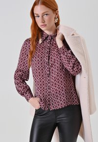 Blusa bordeaux con motivo a disegni geometrici bianchi, davanti con bottoni, maniche lunghe e cappotto beige abbinato, indossata con pantaloni neri.
