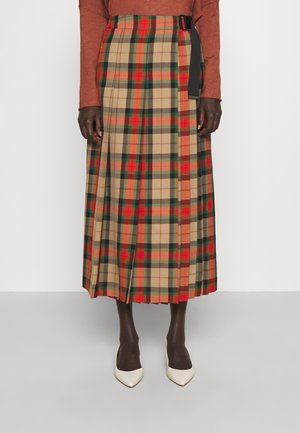 Jupe plissée mi-longue à motif à carreaux beige, rouge, orange et vert, portée avec un haut à manches longues couleur rouille et des chaussures pointues blanches.