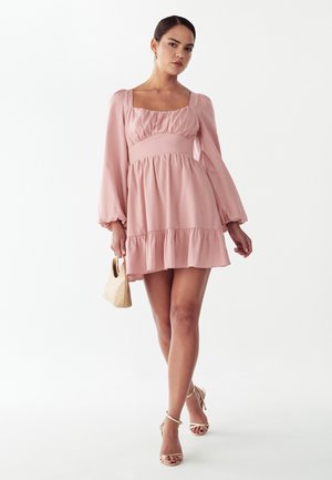 Žena v ružových mini šatách s dlhým rukávom a nafúknutými rukávmi, drží malú tkanú kabelku a kráča v béžových sandáloch na opätku.