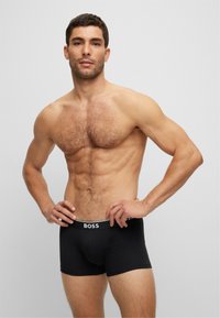 Schwarze Boxer-Briefs mit einem weißen "BOSS"-Bund, aus Baumwollmaterial, figurnaher Schnitt und glatter Textur. Das Modell pose mit den Händen auf den Hüften.