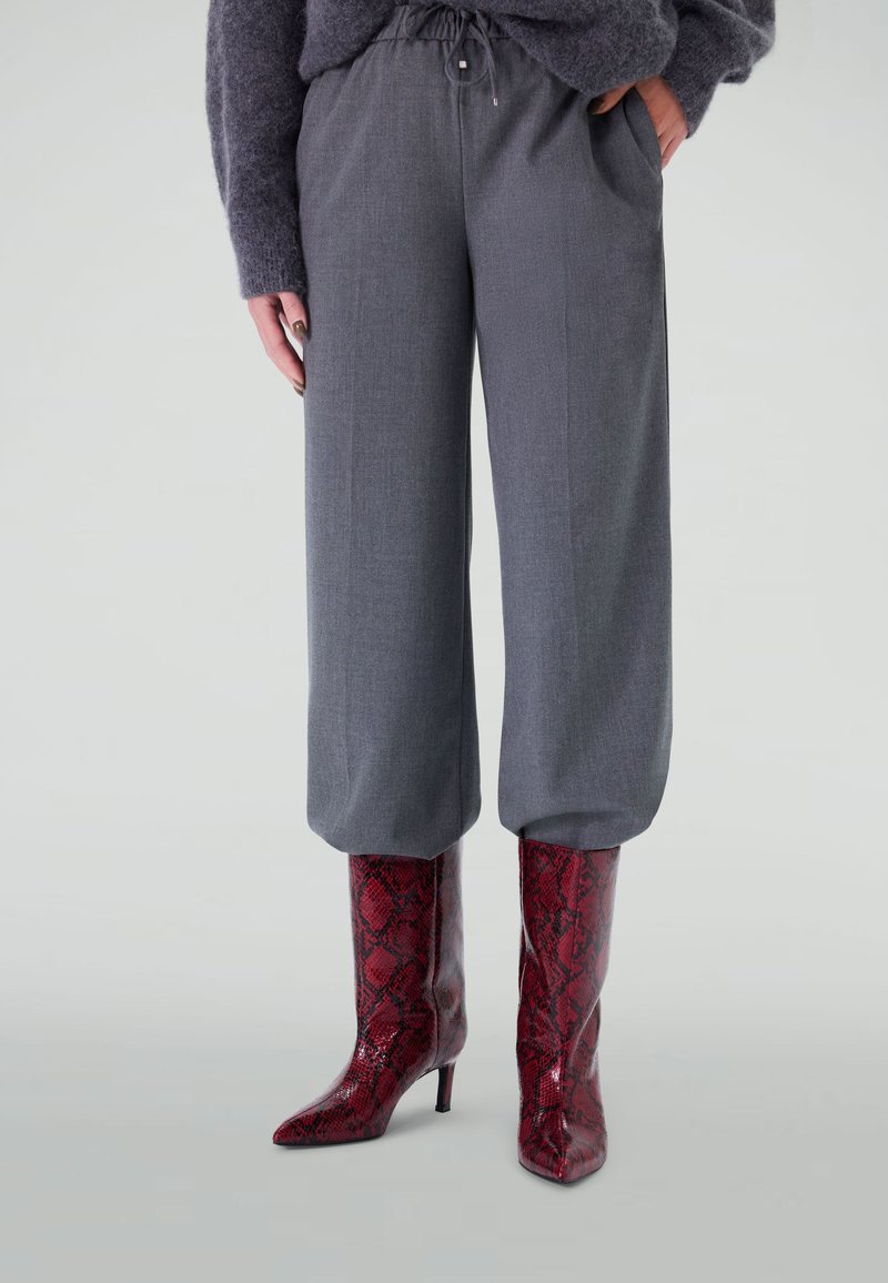 Femme portant un pantalon taille haute gris avec cordon de serrage et des bottes à talons en cuir de serpent rouge sur fond clair.