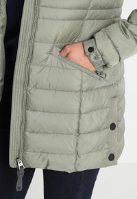 Chaqueta de plumas verde claro con patrón acolchado, bolsillo con cremallera y detalles de botones a presión en los puños y laterales. Textura suave y lisa.