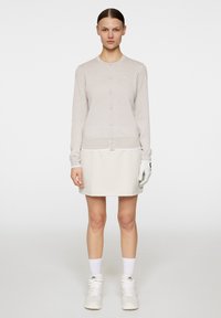 Beige kofta med knappar, i kombination med en ljusbeige kjol. Modellen bär vita sneakers och en golfhandske, framför en enkel bakgrund.
