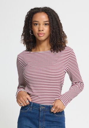 Jeune femme aux cheveux bouclés portant des boucles d'oreilles créoles, un t-shirt à manches longues rayé rouge et blanc, et un jean bleu, debout devant un fond uni.