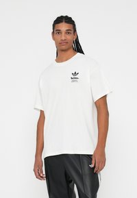 White cotton t-shirt with short sleeves, featuring a black printed logo on the chest. Paired with black leather pants. Simple design.