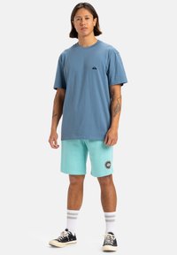 Camiseta de algodón azul claro con un pequeño logo en el pecho, combinada con pantalones cortos verde menta que tienen un parche circular, y zapatillas negras.