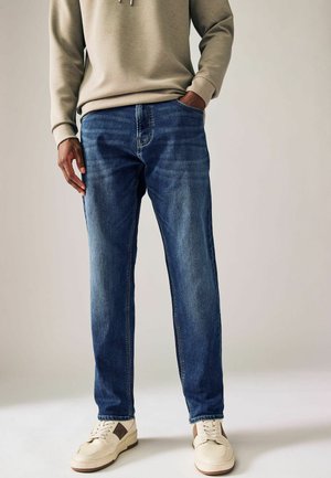 Mid-rise blue denim jeans with a straight leg cut, subtle fading, five pockets, and contrast stitching. Worn with a beige sweatshirt and white sneakers.
