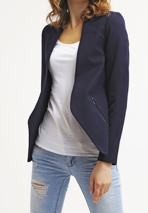 Femme portant un blazer bleu marine taillé sur un t-shirt blanc et un jean bleu clair déchiré, debout devant un fond uni.