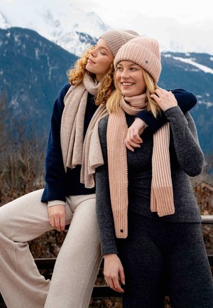 Due persone che indossano morbidi e testurizzati berretti e sciarpe. Un berretto è a righe rosa, l'altro è beige uniforme, abbinati a maglioni scuri e chiari.