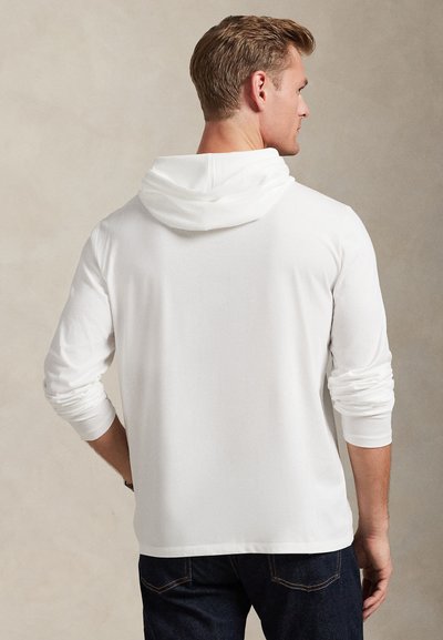 Čovjek okrenut leđima koji nosi dugu bijelu majicu s kapuljačom s kapuljačom spuštenom i tamnoplavim trapericama na jednobojnoj pozadini.