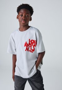 Lysegrå T-shirt med korte ærmer og fed rød tekst "ALLSAINTS LONDON" på forsiden. Har en afslappet pasform og blødt stof.