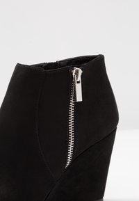 Czarny botek na koturnie z zamszowym wykończeniem, z bocznym zamkiem błyskawicznym i teksturowanymi akcentami w smukłym, zwężającym się fasonie.