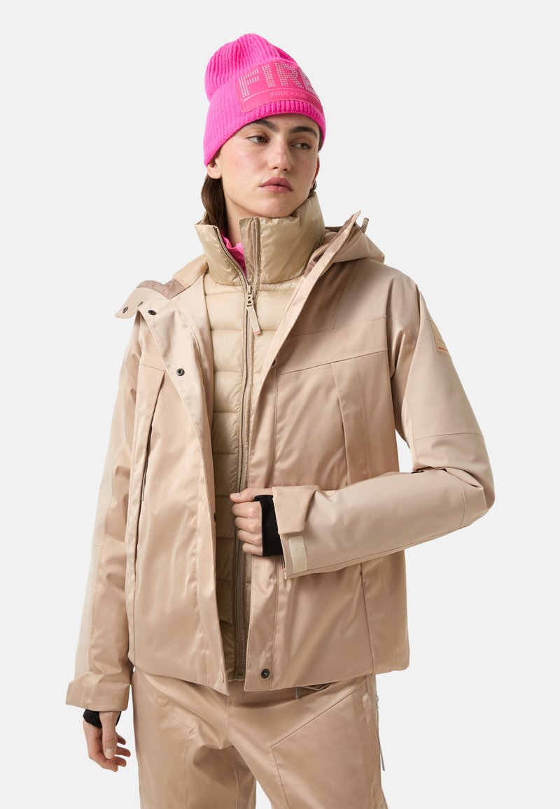 Beige geïsoleerd jack met een gladde textuur, ritssluiting en drukknoopsluiting, met zakken, gedragen over een lichter beige binnenjack. Roze gebreide muts.