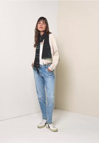 Casual outfit met een crème bomberjack, zwart gestreepte sjaal, witte top, lichtblauwe jeans en witte sneakers met groene accenten.