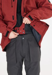 Rote wasserfeste Jacke mit schwarzem Innenfutter und schwarzem Reißverschluss, kombiniert mit grauen Hosen, die eine strukturierte Taille und große Seitentaschen haben.
