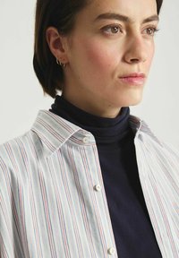 Chemise à rayures en tissu léger avec des rayures verticales bleu, rouge et blanc, portée sur un col roulé sombre. Boucles d'oreilles en or visibles.