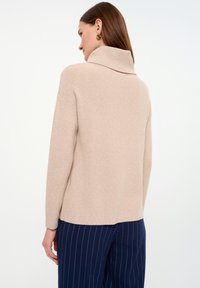 Beige ribbed sweater with a high collar, featuring a relaxed fit. The model wears navy striped pants, showcasing a smooth, neutral-colored texture.