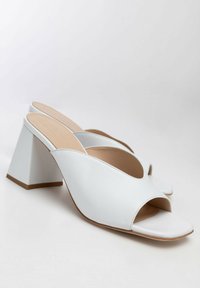 Mule bianche in pelle aperte sulla punta con ampi tacchi a blocco e design scorrevole, esposte su uno sfondo bianco uniforme.