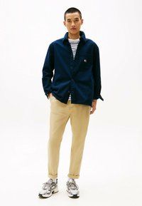 Camicia blu navy con bottoni e tasca sul petto, indossata sopra una maglietta a righe. Pantaloni beige e scarpe sportive completano l'outfit. Design minimalista.