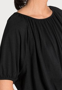 Sort top med rund halsudskæring og samlede detaljer omkring skuldrene. Stoffet er blødt med et løs, flydende design.