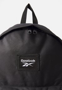 Schwarzer Rucksack aus robustem Material mit runder Form, Oberhandgriff und doppeltem Reißverschluss. Enthält ein auffälliges Reebok-Logo-Patch.