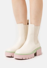 Jasnokremowe botki za kostkę o gładkiej skórzanej fakturze, z masywnymi różowymi podeszwami i zielonymi akcentami, zaprojektowane z elastycznymi bocznymi panelami.