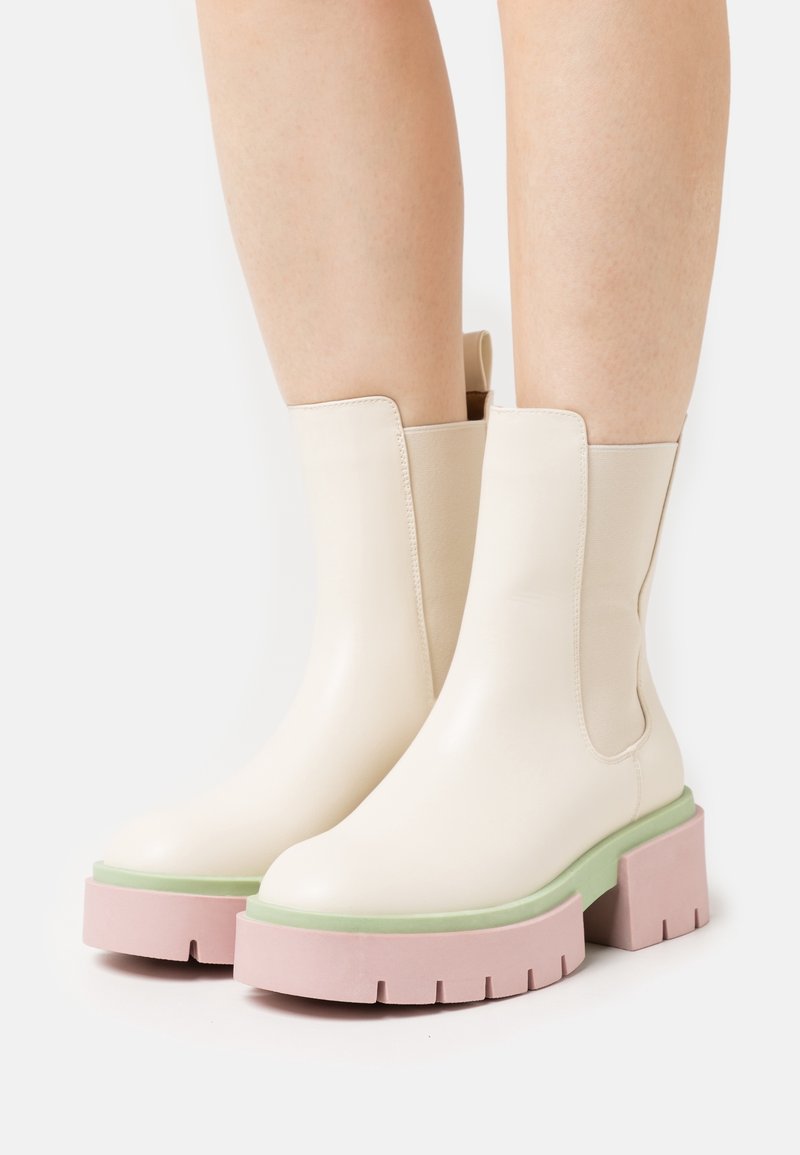 Jasnokremowe botki za kostkę o gładkiej skórzanej fakturze, z masywnymi różowymi podeszwami i zielonymi akcentami, zaprojektowane z elastycznymi bocznymi panelami.