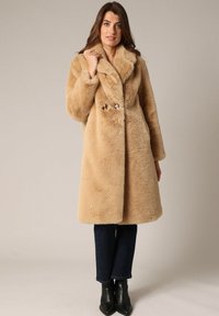 Abrigo de piel sintética beige con un amplio cuello, cierre de doble botón y mangas largas. Lleva puesto sobre unos vaqueros oscuros y botas negras de tobillo.