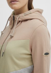 Hooded sweatshirt in light pink and green with a zip closure, featuring braided drawstrings and a logo patch on the sleeve. Soft fabric.