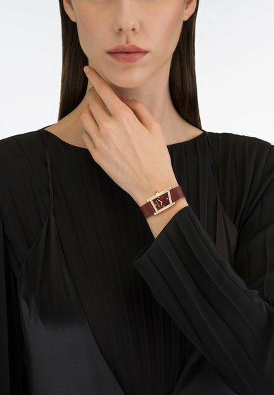 Burgundska usnjena ura z rektangularno ohišje v zlati barvi, ki ima minimalistično številčnico in gladek trak. Nosila se je na črnem nabranem rokavu.