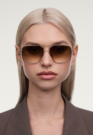 Braune Verlaufssonnenbrille mit einem Metallrahmen, die eine rechteckige Form und dünne silberne Akzente aufweist. Tragen von einem Modell vor einem neutralen Hintergrund.